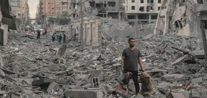 Man walking through neighborhood destroyed by bombs, holding a large stuffed teddy bear and Minnie Mouse